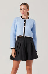 A woman is wearing a light blue cropped cardigan with black trim and large decorative buttons down the front. The cardigan has long sleeves, and the black trim is present along the collar, cuffs, and hem. She is also wearing a high-waisted black pleated skirt that reaches above her knees.